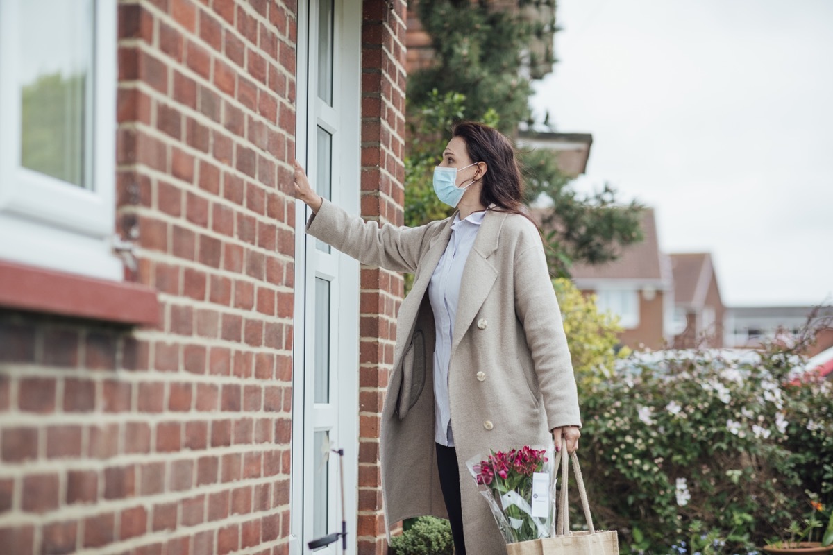 A side view of a female healthcare worker wearing a protective mask knocking on her grandmother's front door, she is dropping off groceries and flowers during the Coronavirus pandemic.