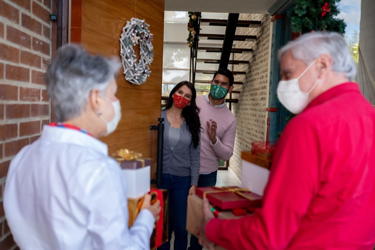 Happy senior couple arriving home for Christmas wearing facemasks and carrying presents while greeting to their kids - COVID-19 pandemic lifestyle concepts