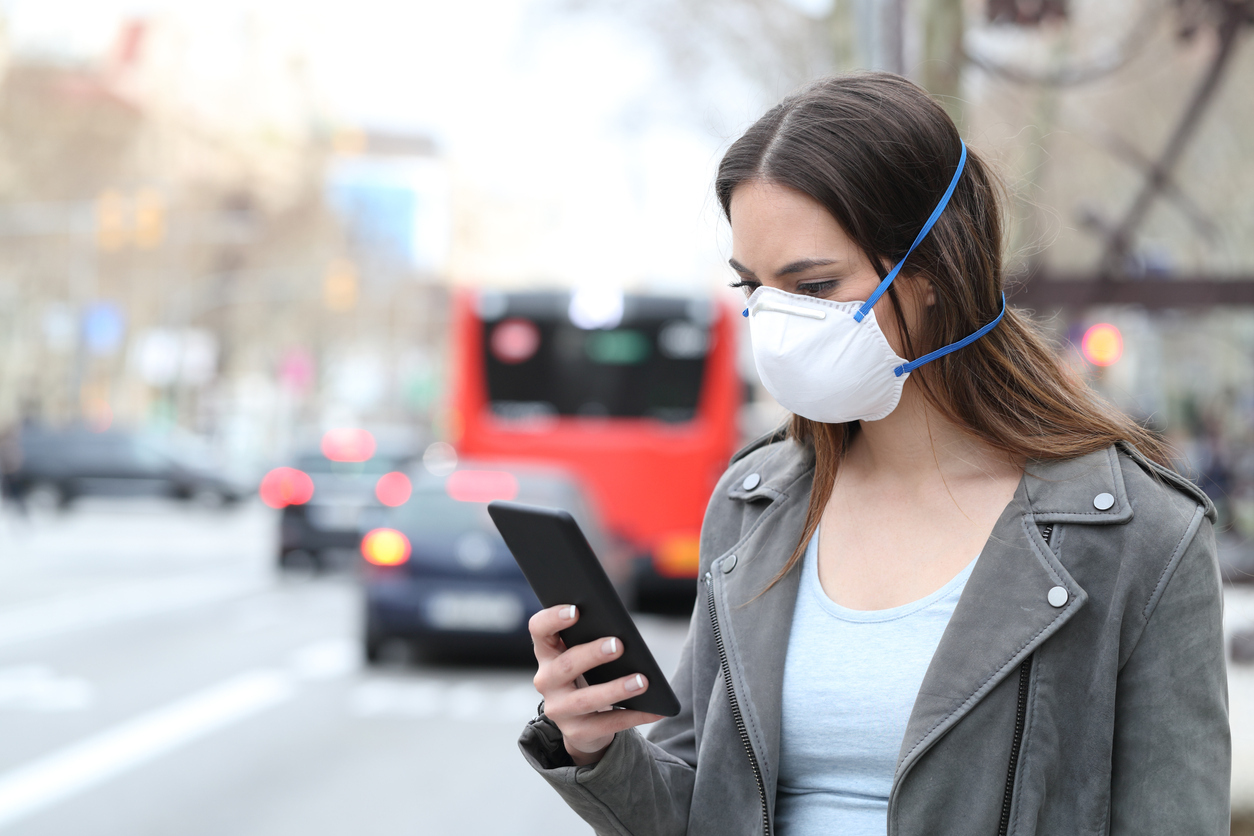A young woman standing on the sidewalk of a city wearing a face mask checks her smartphone.