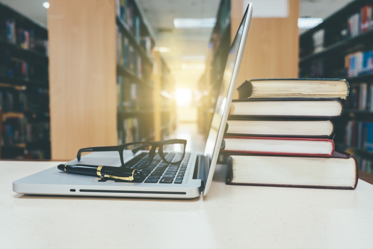 A laptop, a stack of books, a pen, and some eyeglasses sitting on a table in a library