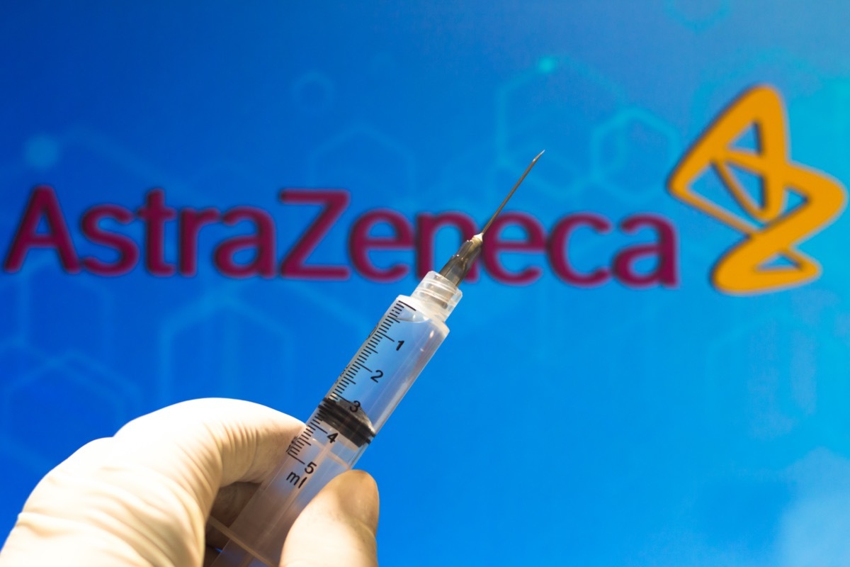 A medical syringe (coronavirus vaccine) is seen with AstraZeneca PLC company logo displayed on a screen in the background.