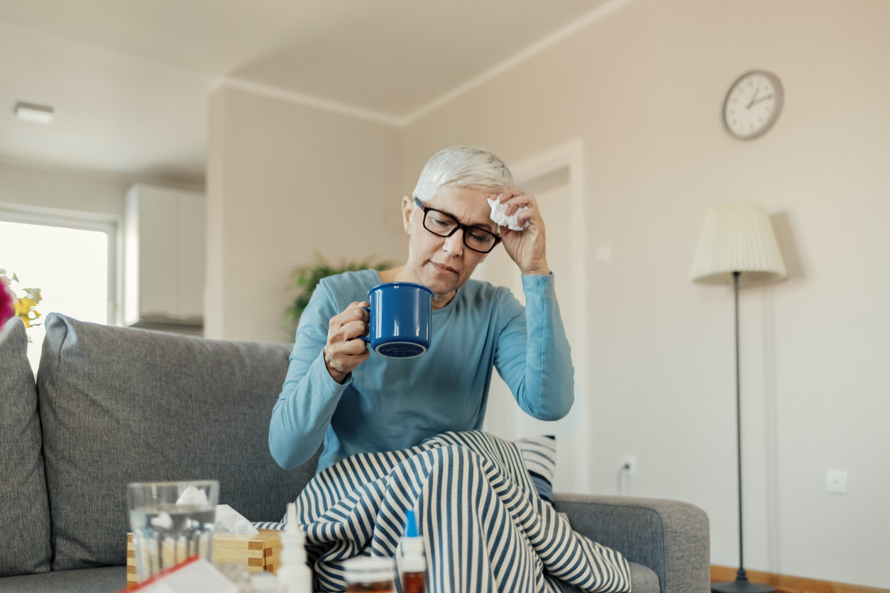 An older woman sits on the couch with symptoms of the common cold, holding a mug and tissue