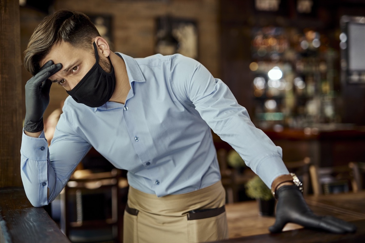 Distraught waiter holding his head in pain while wearing protective face mask and working in a pub.