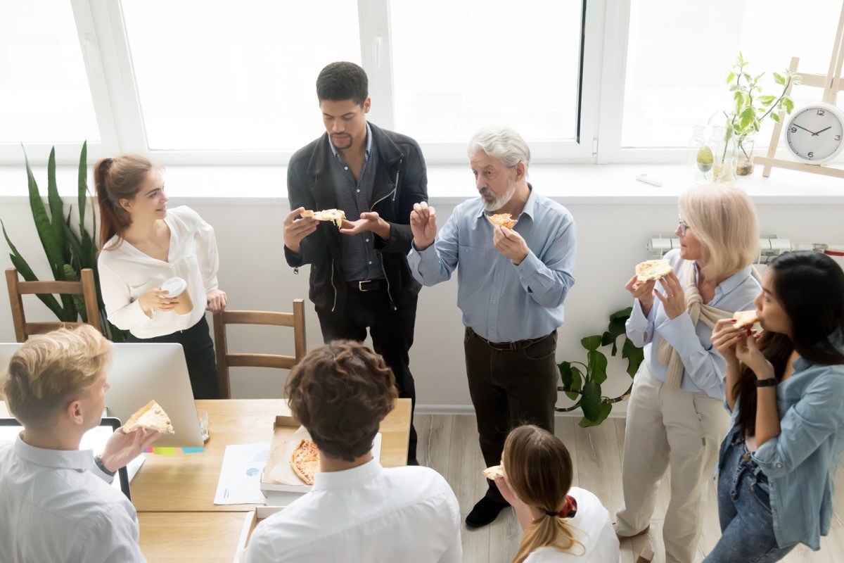 men and women standing and eating pizza in office