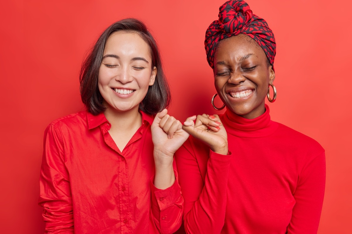 Happy positive women hook each others little fingers in conciliation or friendship smile toothily keep eyes closed stand closely against bright red background wear casual clothes. Relationship concept