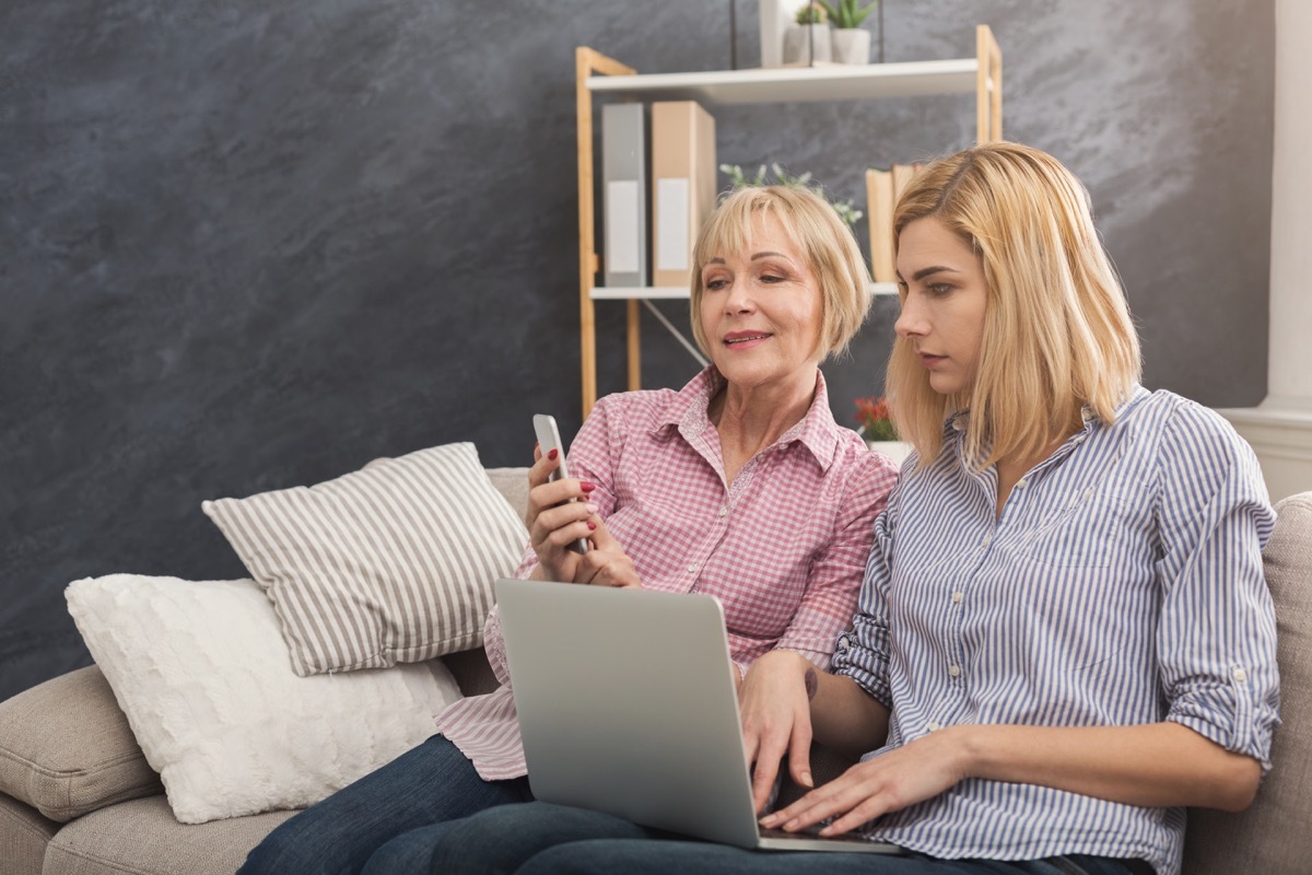 Happy adult mother holding smartphone and daughter using laptop, reading news at home. Woman pointing on screen. Generation and relationship concept