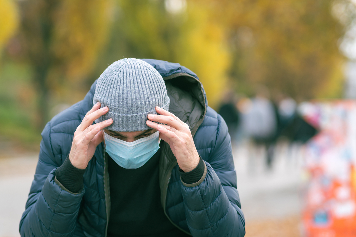 A man in a medical mask in the park with his hand on his head due to a headache.