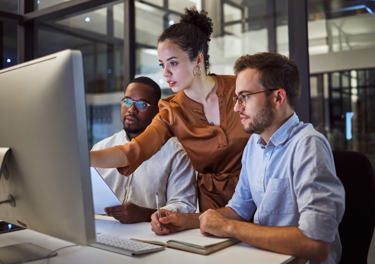 two men sitting at computers with woman standing between them pointing at something on screen