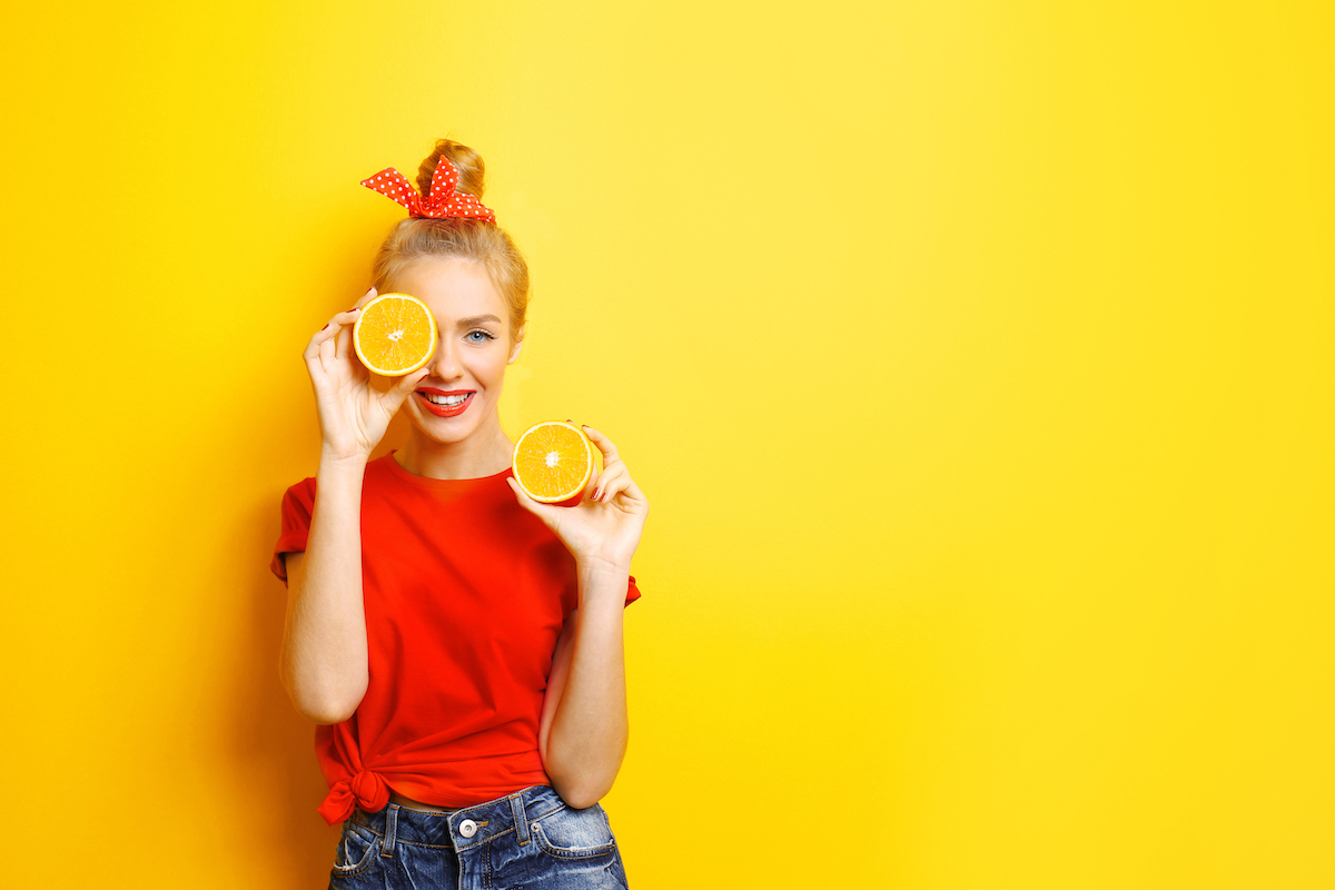 A young woman in a red t-shirt with her blonde hair tied up in a red bow, holding two orange slices against a yellow background.