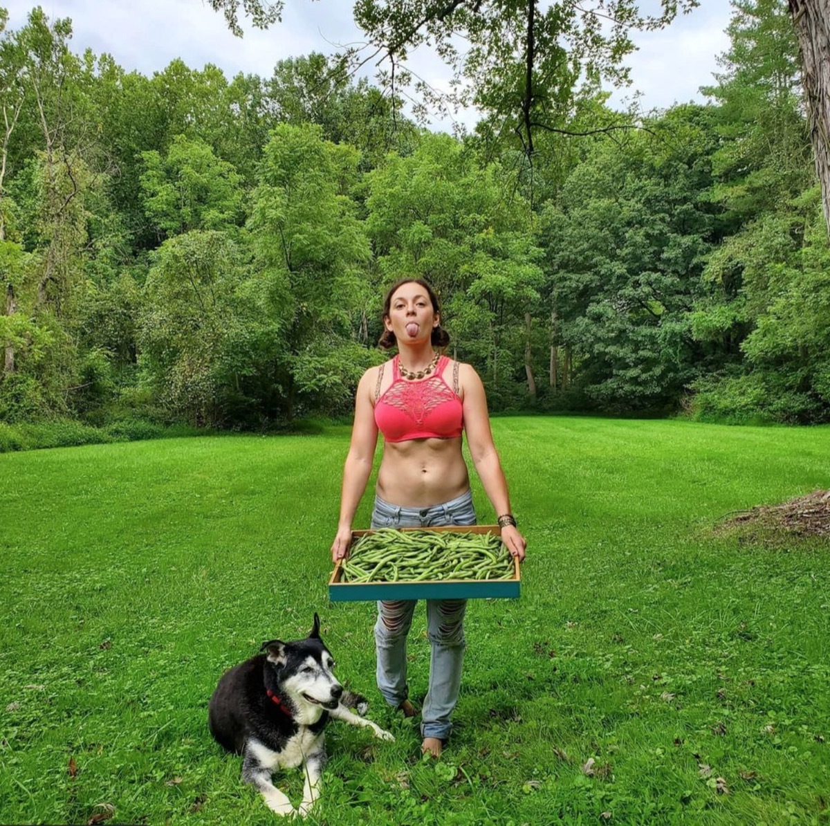 Mackenzie Rosman wearing jeans and a red crop top in a green field carrying a tray of vegetables and standing next to a black and white dog