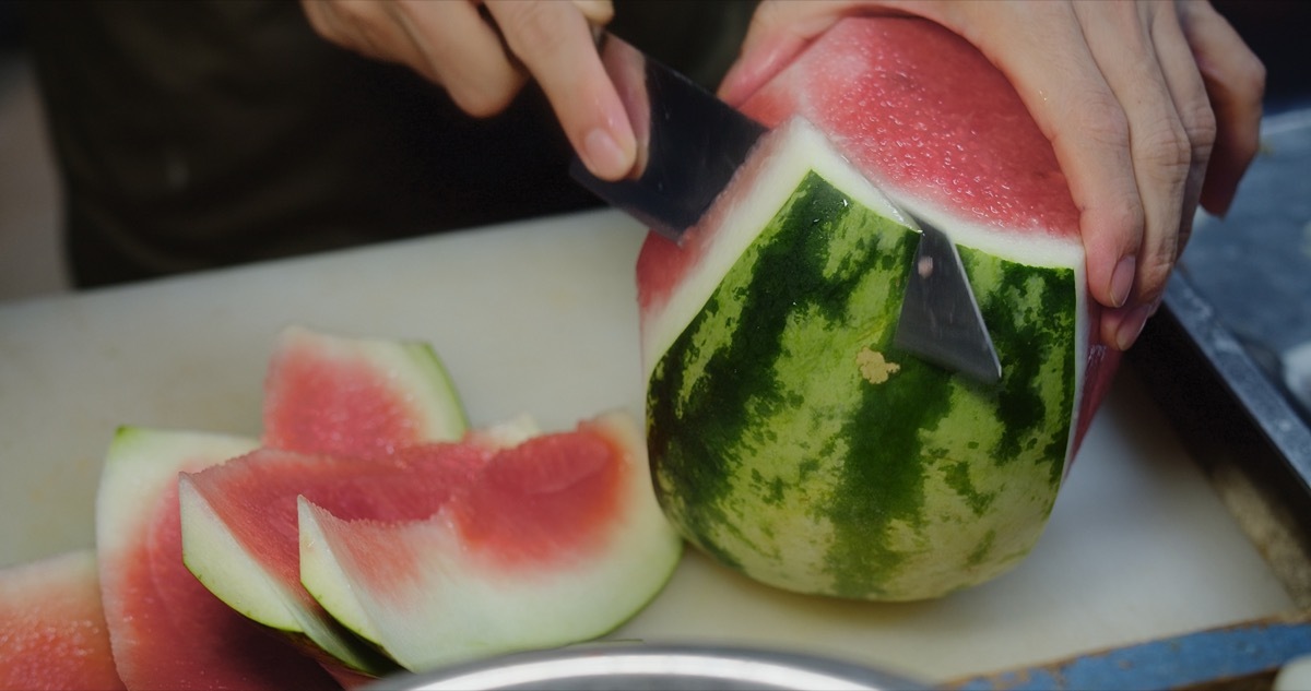 Close-up of hands cutting a fresh watermelon on a kitchen countertop, slices ready to serve.