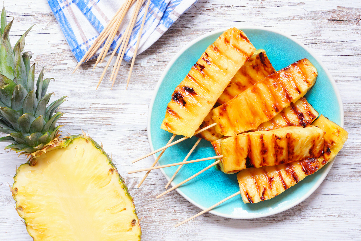 top-down view of grilled pineapple skewers and a half a pineapple on a white wood background