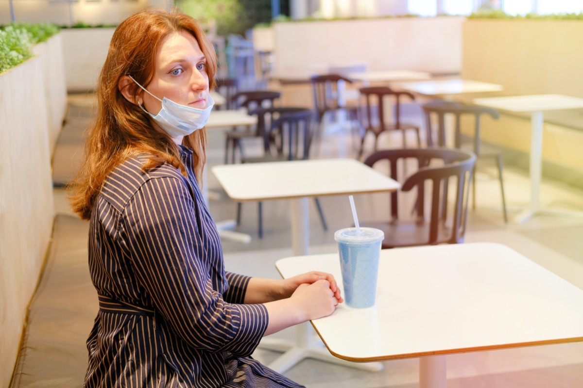 Young redhead woman with a medical mask removed sits in an empty cafe after quarantine. Deserted restaurant after the completion of the coronavirus epidemic