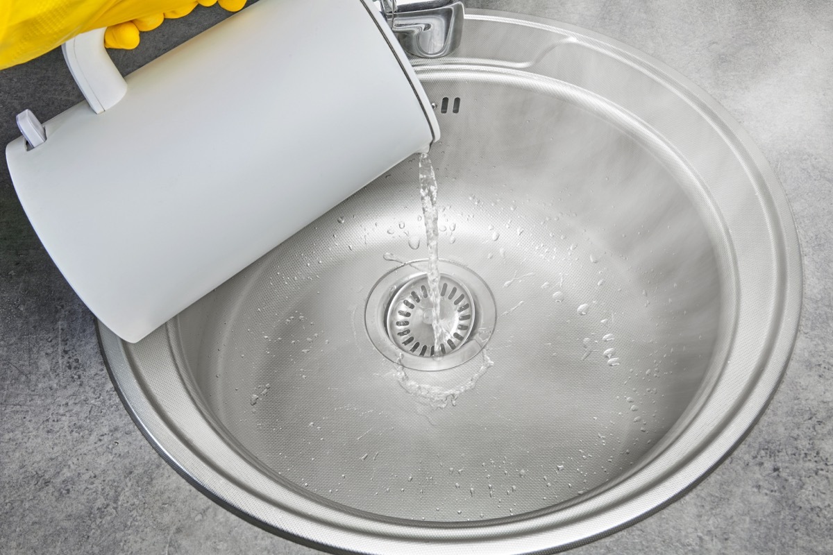 A person in yellow rubber gloves pours hot water with steam from an electric kettle into a metal sink. Cleaning and disinfection of sanitary ware in the kitchen