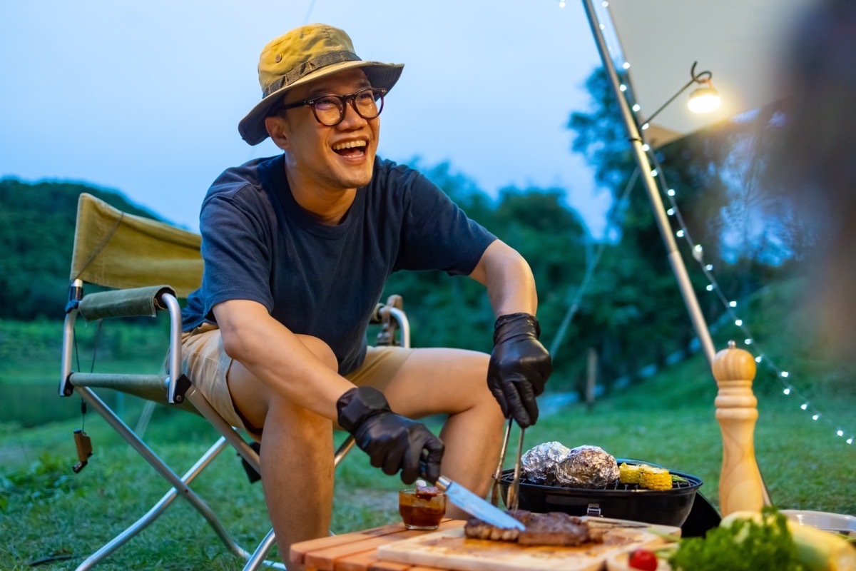 Asian man cooking meal grilled bbq steak for dinner party at camp at sunset. Group of People enjoy outdoor adventure lifestyle travel nature hiking and camping together on summer holiday vacation.