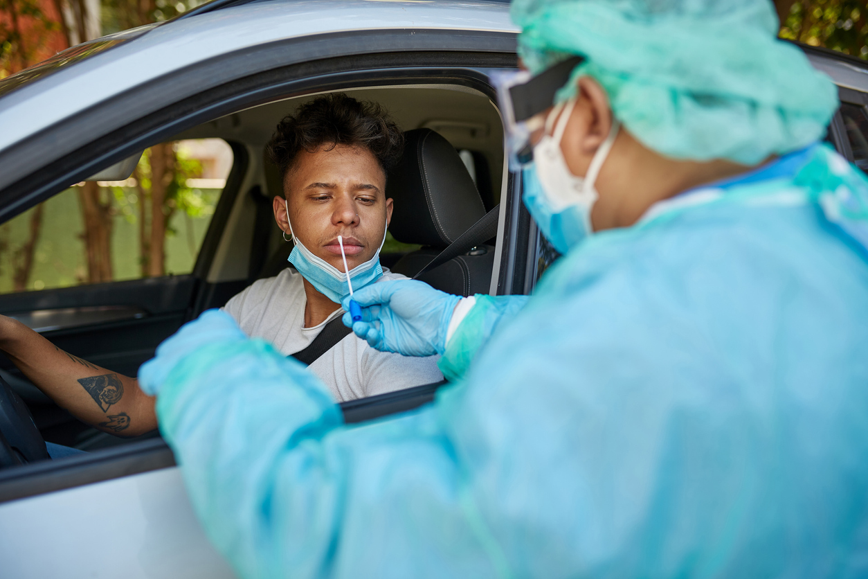 A health care worker in full protective gear performs a nasal swab on a young man sitting in his car to test for coronavirus