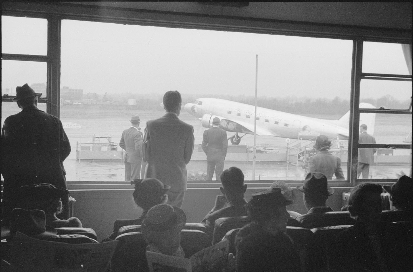 a group of travelers wait in an airport gate and watch out the windows to the runway