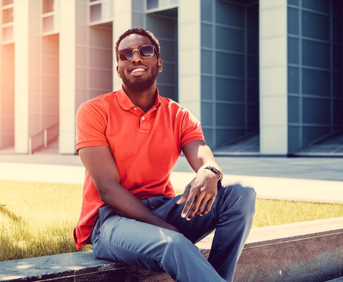 A positive Black male dressed in a red polo shirt posing on a street in a downtown. - Image