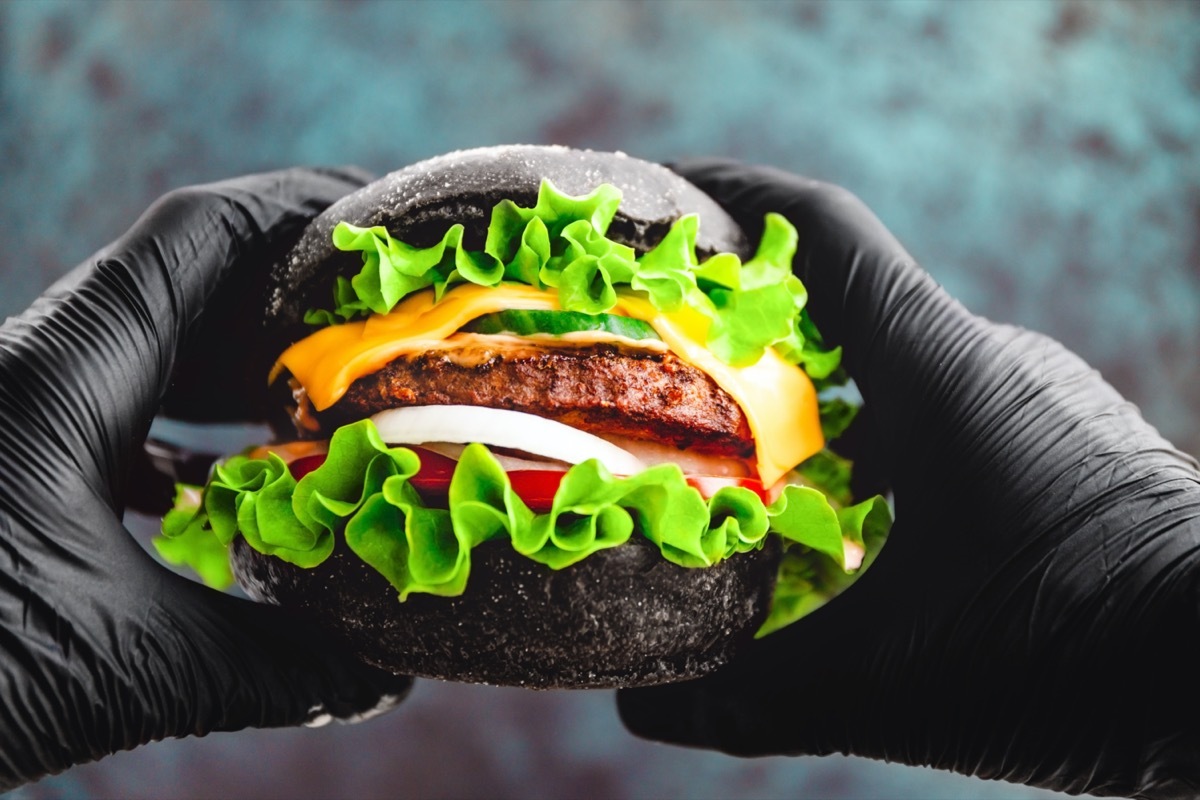 Hands in black gloves hold a big black burger with marble beef patty, cheese and fresh vegetables.