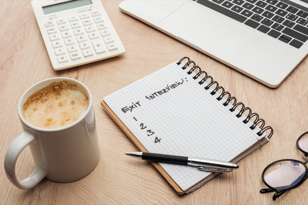 Notebook with exit interview lettering and numbers on wooden table with laptop, stationery, glasses and cup of coffee