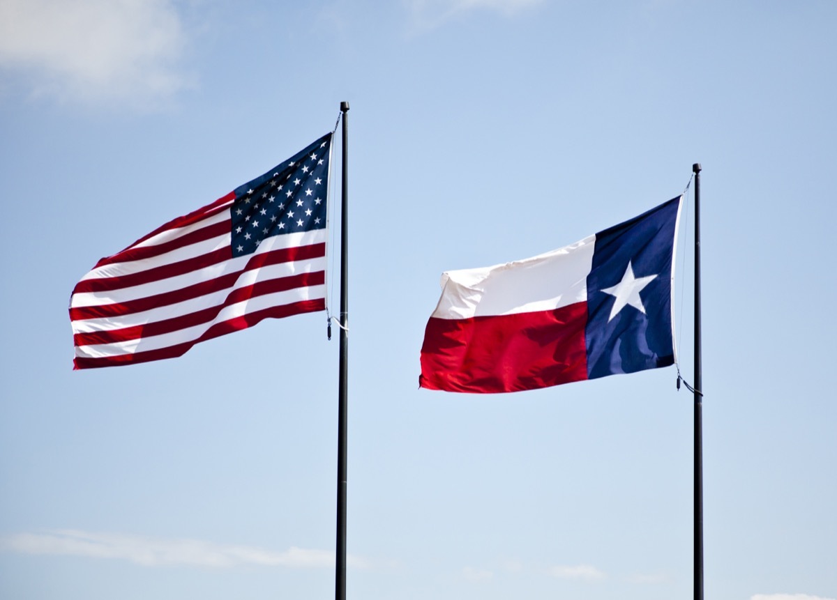 The American and Texas flags flying high together against a blue lightly cloudy sky