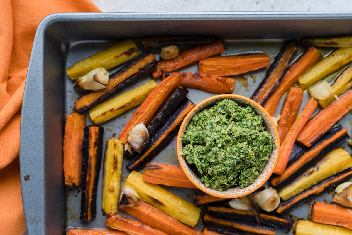 Top view and close up of a baking/frying sheet with roasted/baked rainbow orange, yellow and purple carrots with garlic served with basil, parmesan and olive oil green pesto with an orange napkin.