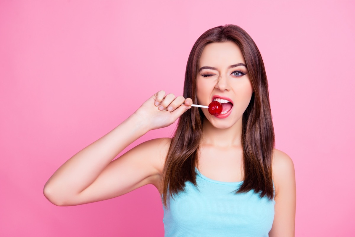 woman with straight brown hair trying to bite red lollipop on stick