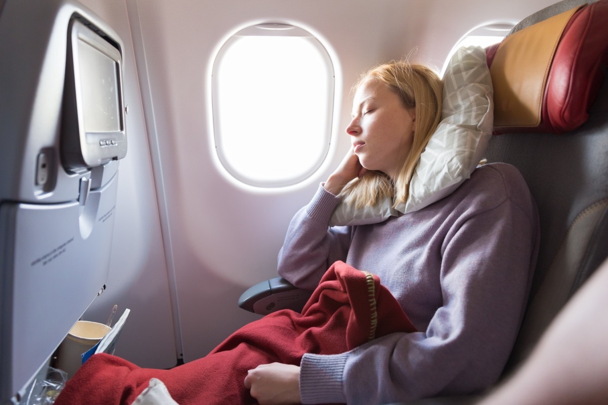 young blonde woman sleeping on a plane, using a neck pillow and leaning against the window