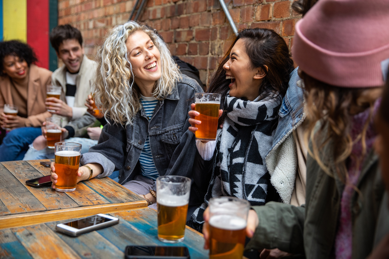 A group of young friends laughing and enjoying beers at a pub while not wearing face masks.