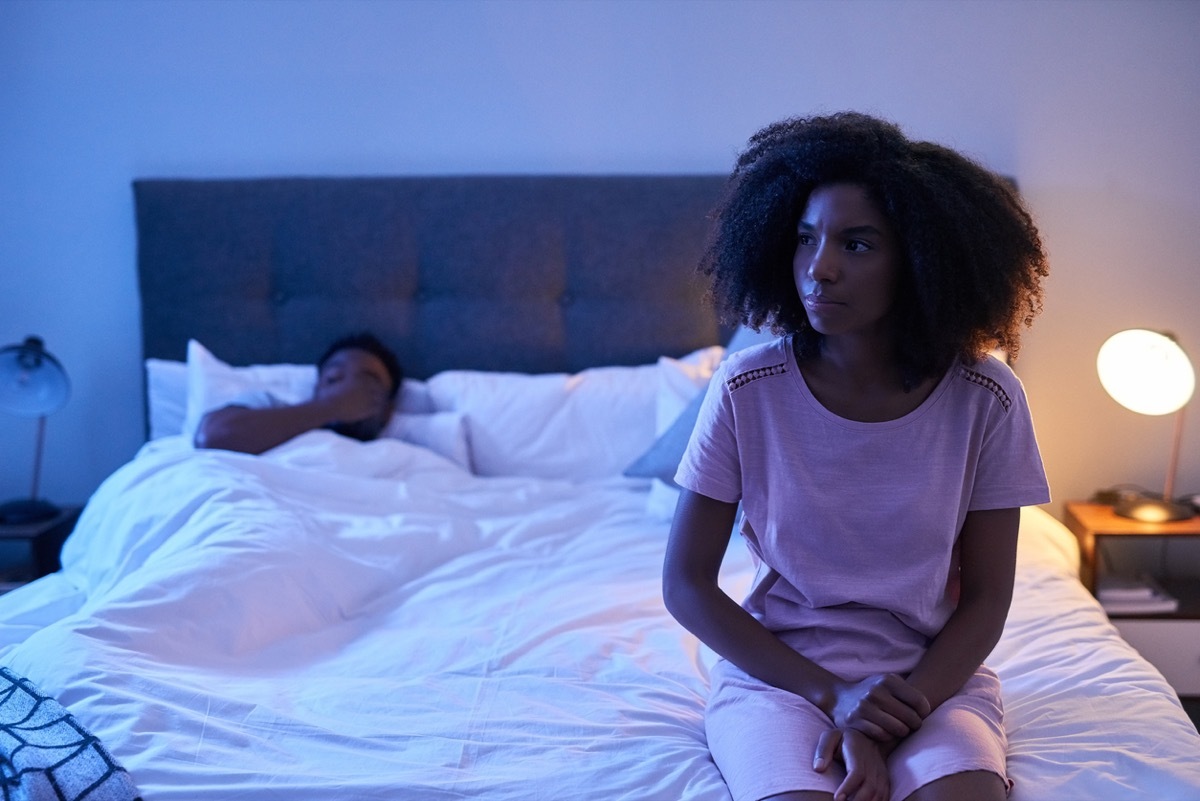 young black woman sitting up worried in bed with partner in the background