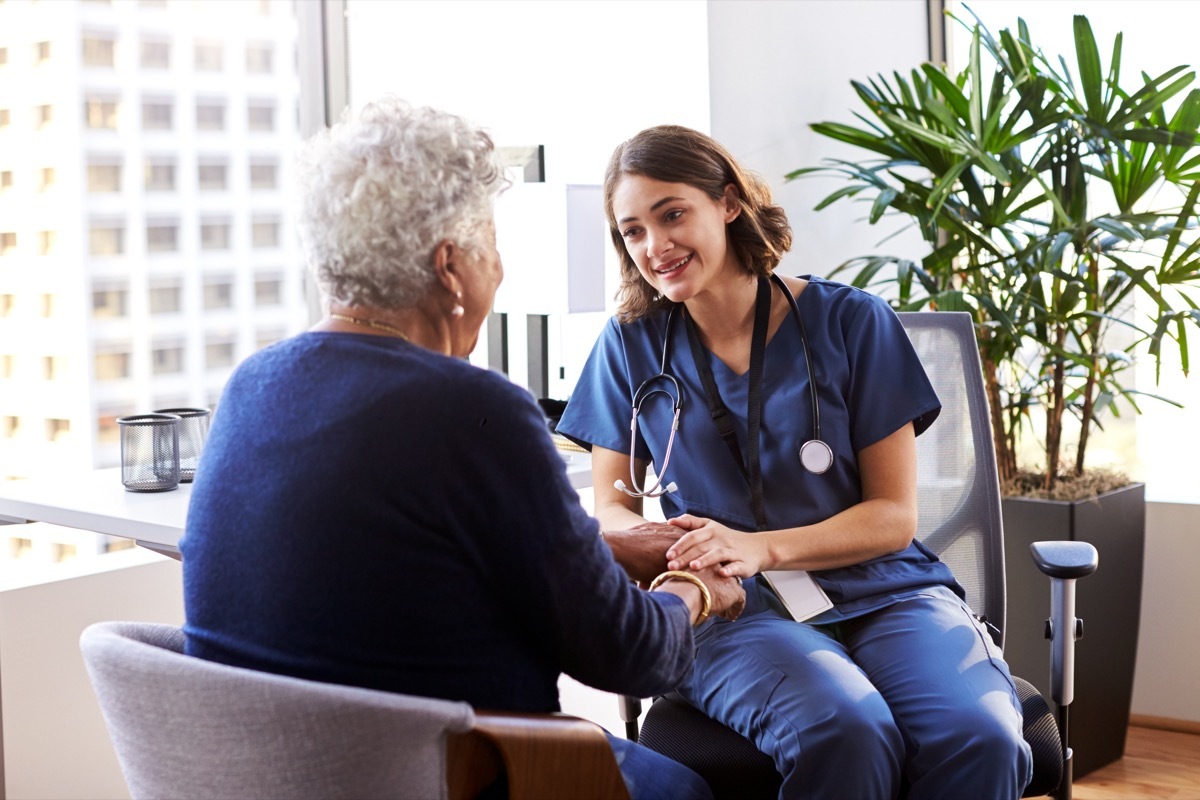 Nurse Wearing Scrubs In Office Reassuring Senior Female Patient And Holding Her Hands