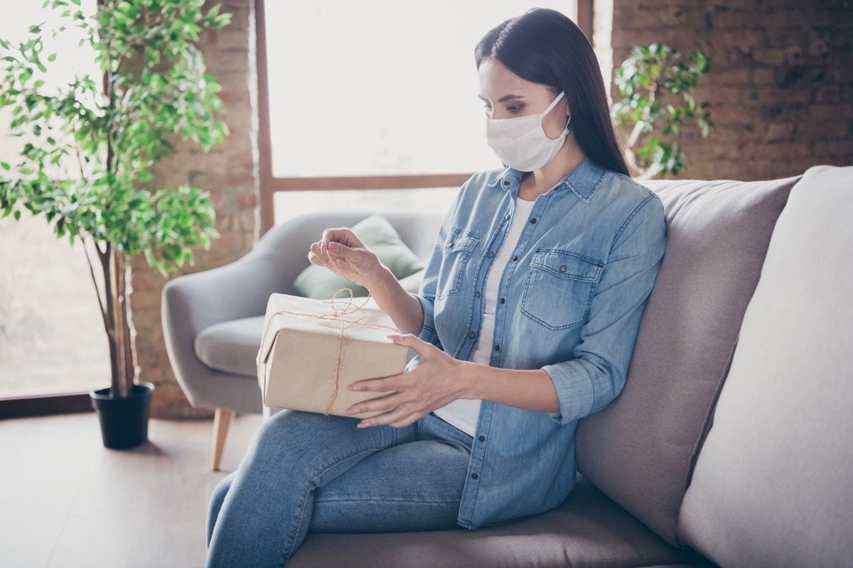 woman wearing mask opening package on couch inside home