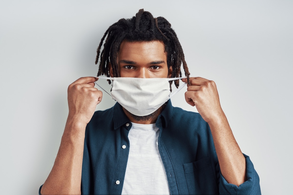 Charming young man adjusting medical face mask and looking at camera while standing against grey background
