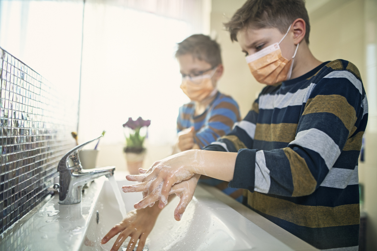 Two young boys wearing face masks wash their hands at a sink.
