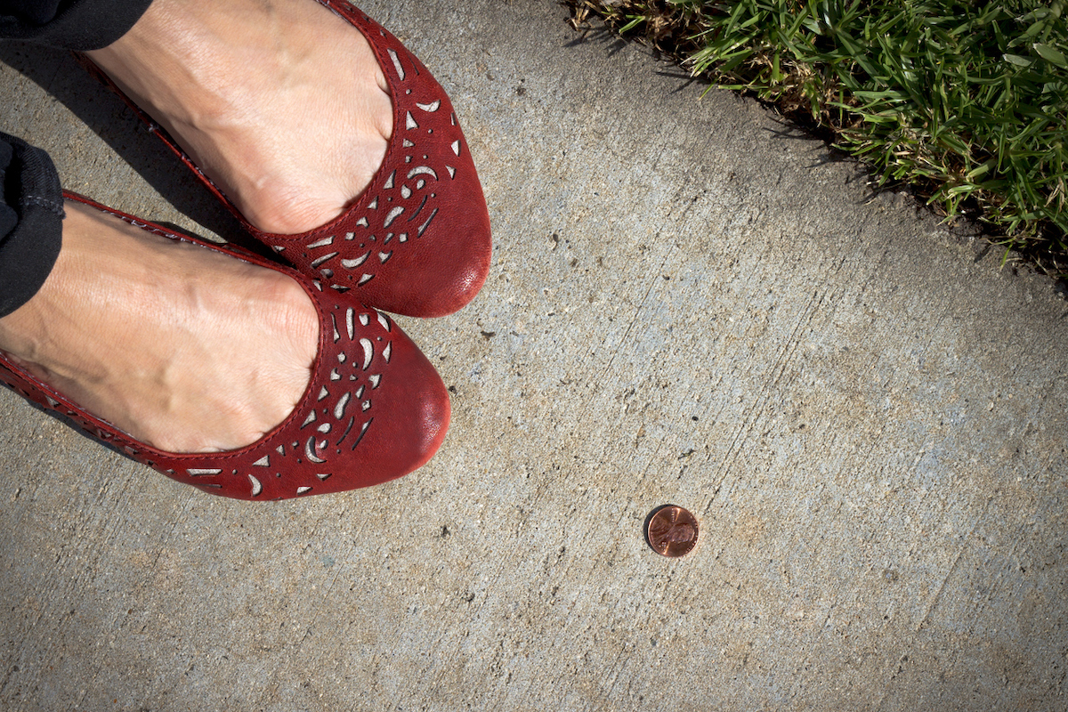 Close up of a women in red flat shoes with a lucky heads-up penny near her feet on the sidewalk.