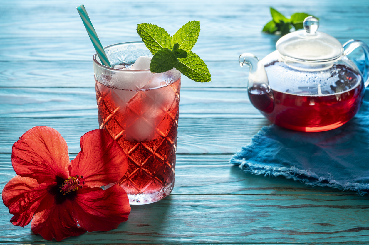 A hibiscus tea cocktail in a glass next to a clear kettle
