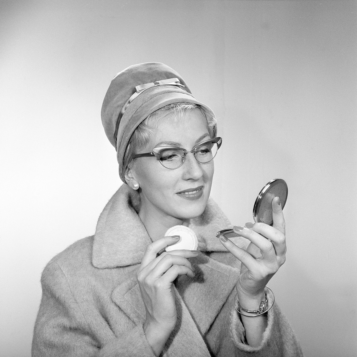 1950s makeup. A young woman is looking at herself in her pocket mirror and improves on her makeup. She is wearing a fashionable hat, typical 50s glasses and a coat. 1950s