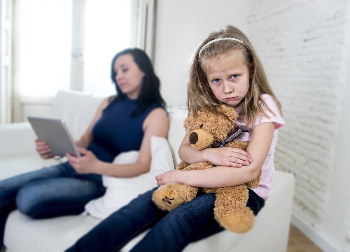 unhappy child sitting on couch while her mother is on her ipad