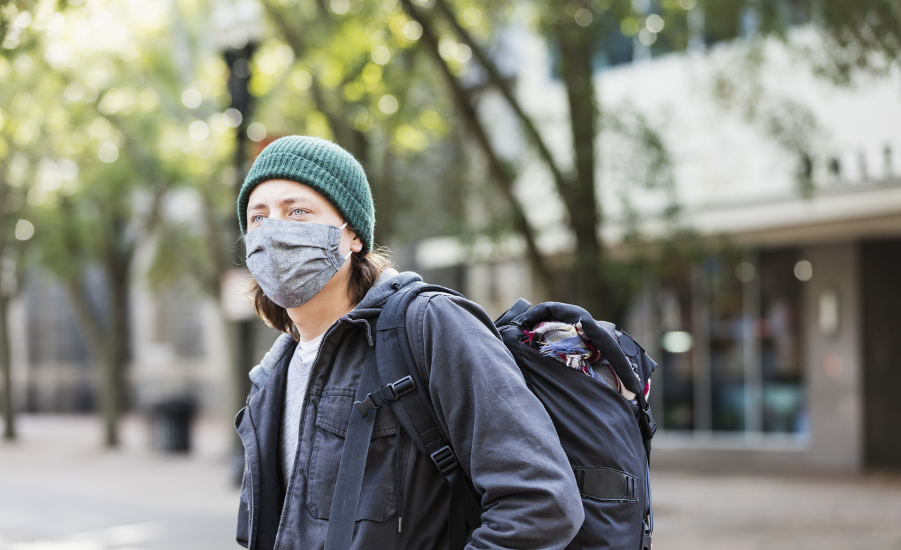 A young man wearing a face mask and a backpack stands on a city street.