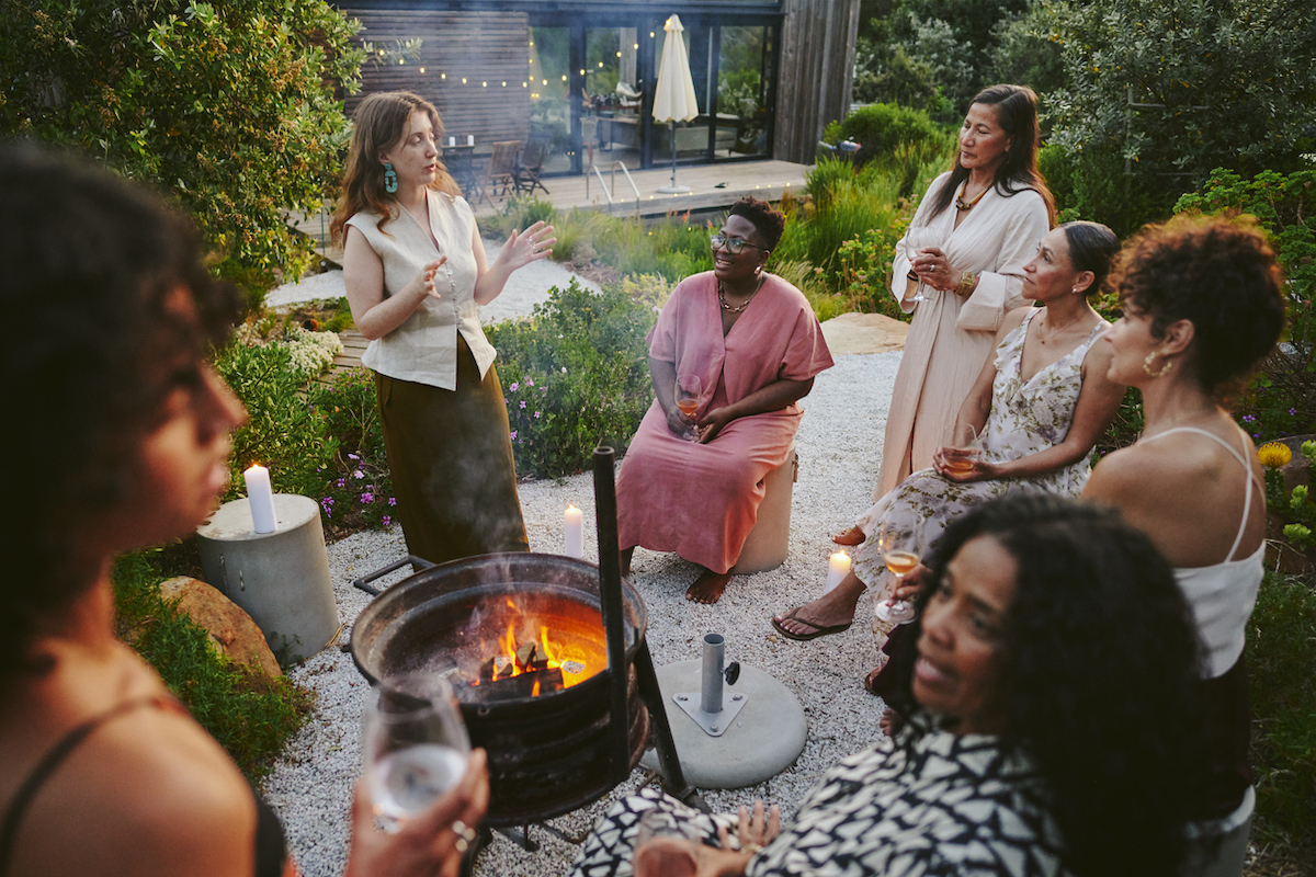 group of smiling women having drinks and talking around a fire pit during an early evening party in the lush garden of a home at dusk