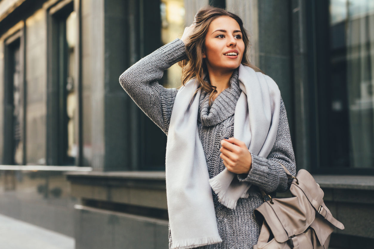 A stylish young woman standing on the street wearing a scarf and grey sweater.