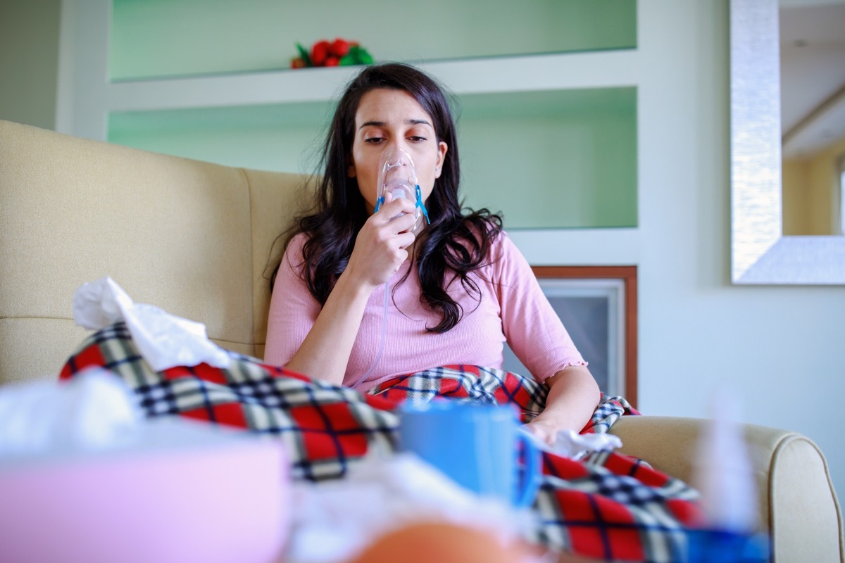 A young woman is suffering from problems with the respiratory system. A beautiful woman with long hair is suffering from pneumonia and using a smoke inhaler at home while sitting on the sofa-COVID-19, (A young woman is suffering from problems with the