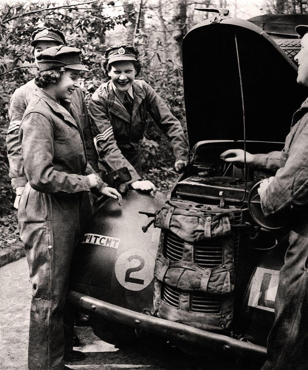 Princess Elizabeth in her uniform of the ATS The Auxiliary Territorial Service, a women's army auxiliary branch, with other smiling ATS service members working on and servicing a British Army military vehicle.