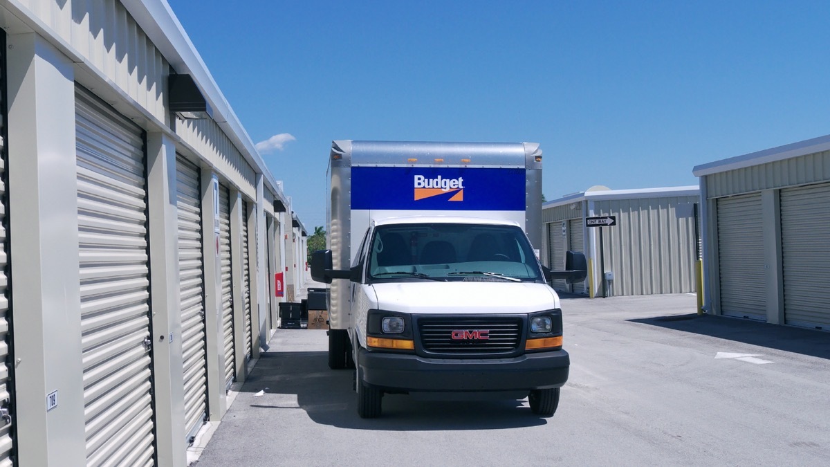 A Budget rental truck is parked on a street between buildings housing self storage units. Budget is a large vehicle rental company in the USA.