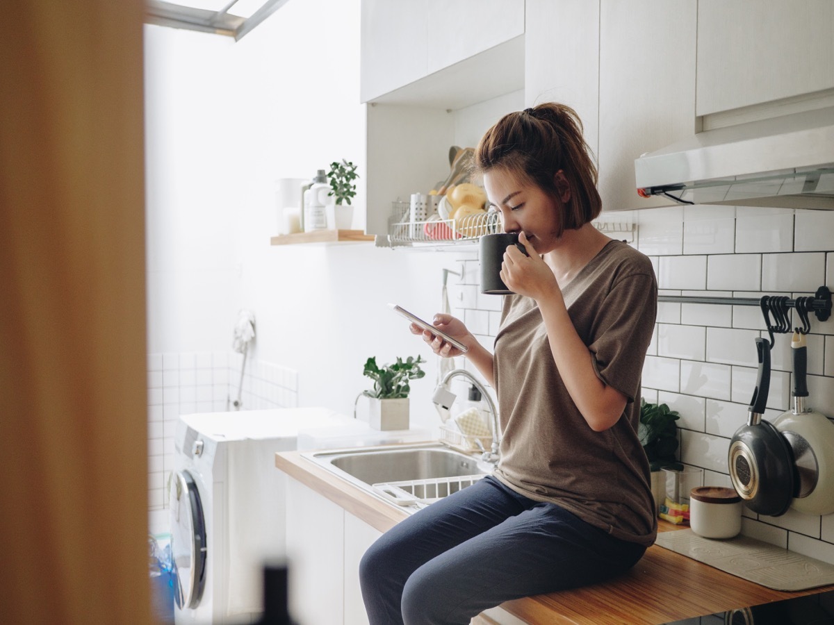 woman drinking coffee while sitting on kitchen counter and working on smart phone in morning at home.