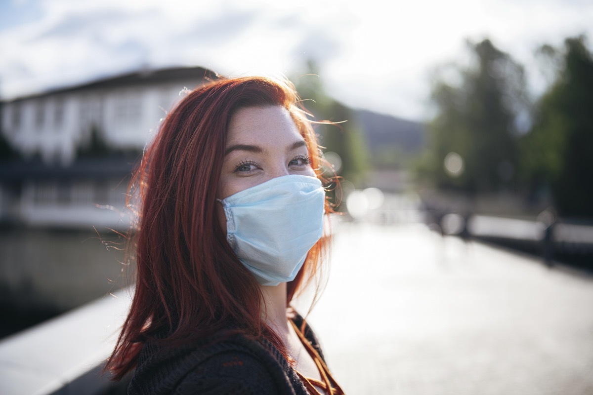 Portrait of woman with red hair wearing protective face mask outdoor and smiling in camera, looking at camera.