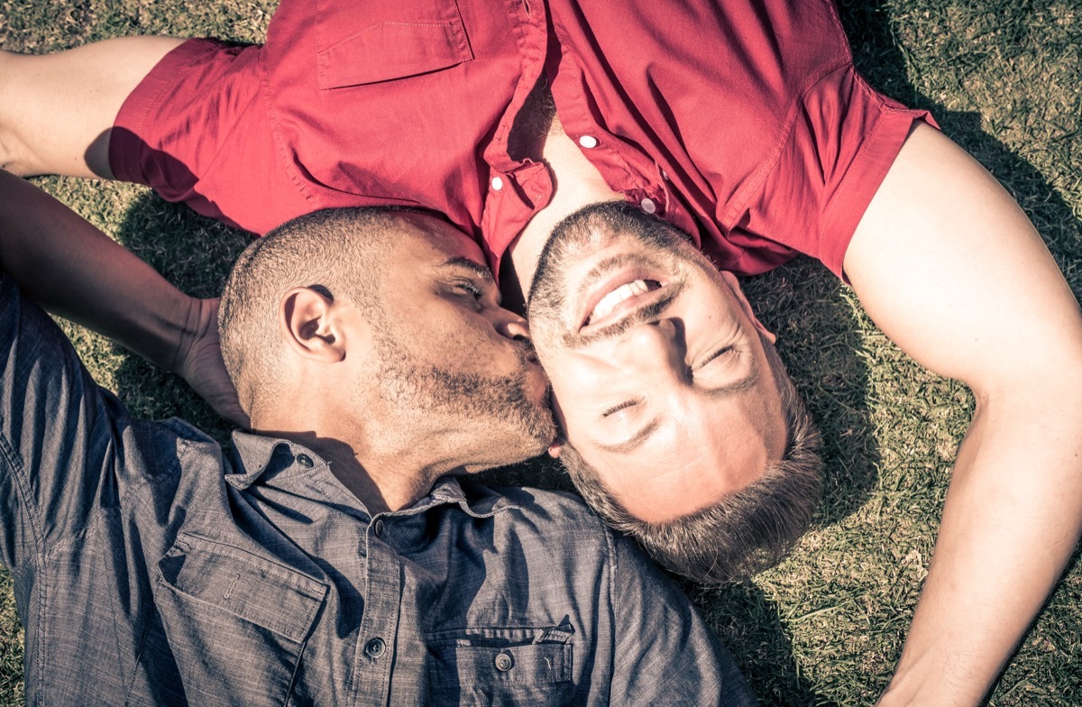 Multicultural gay couple laying in grass kiss on check