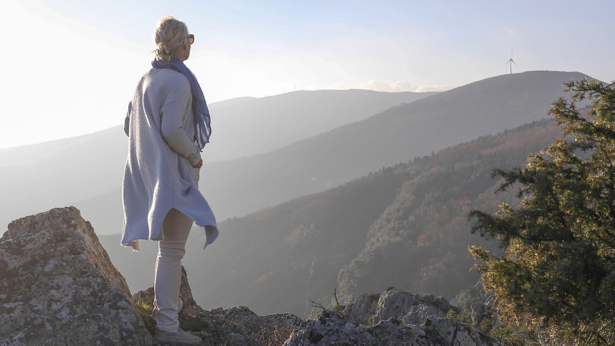 Woman wearing a long sweater and scarf standing on a mountain ridge looking off into the distance.