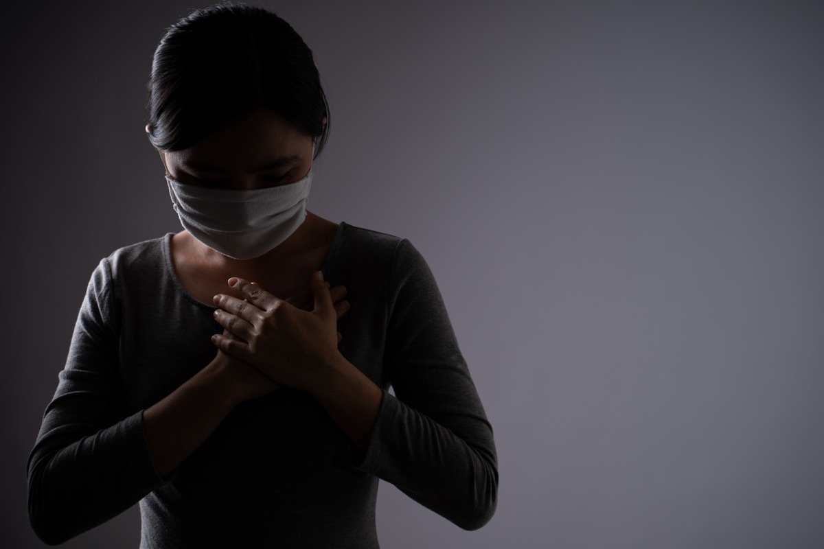woman wearing protective face mask was sick with chest pain, putting her hands on her chest and standing isolated on background. Low key.