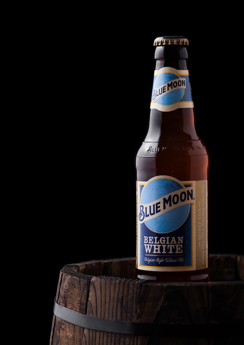 Bottle of Blue Moon belgian white beer, brewed by MillerCoors on old wooden barrel on black background.
